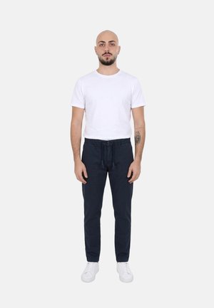 Uomo con testa rasata e barba, indossa una maglietta bianca, pantaloni scuri e scarpe da ginnastica bianche, in piedi con le braccia lungo i fianchi contro uno sfondo semplice.