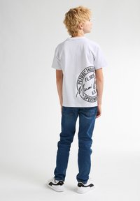 Bílý tričko s krátkým rukávem a černým grafickým potiskem na zadní straně, spárované s modrými džínami a bílými teniskami s černými akcenty.