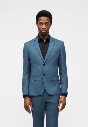 Man standing, wearing a fitted blue suit with a black shirt, against a plain light gray background, facing forward with a neutral expression.