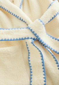 Viltojuma kažokādas halāts krēmkrāsā ar zilu viļņotu apmali. Mīksta tekstūra, krustota dizains ar sasienamu priekšpusi, izceļot mīkstu audumu un kontrastējošus akcentus.