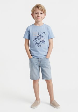 Ξανθός αγόρι με ανοιχτό μπλε μπλουζάκι με τύπωμα σκελετού δεινόσαυρου, ανοιχτόχρωμα τζιν σορτς και μπεζ παπούτσια χωρίς κορδόνια, στέκεται με τα χέρια στις τσέπες.