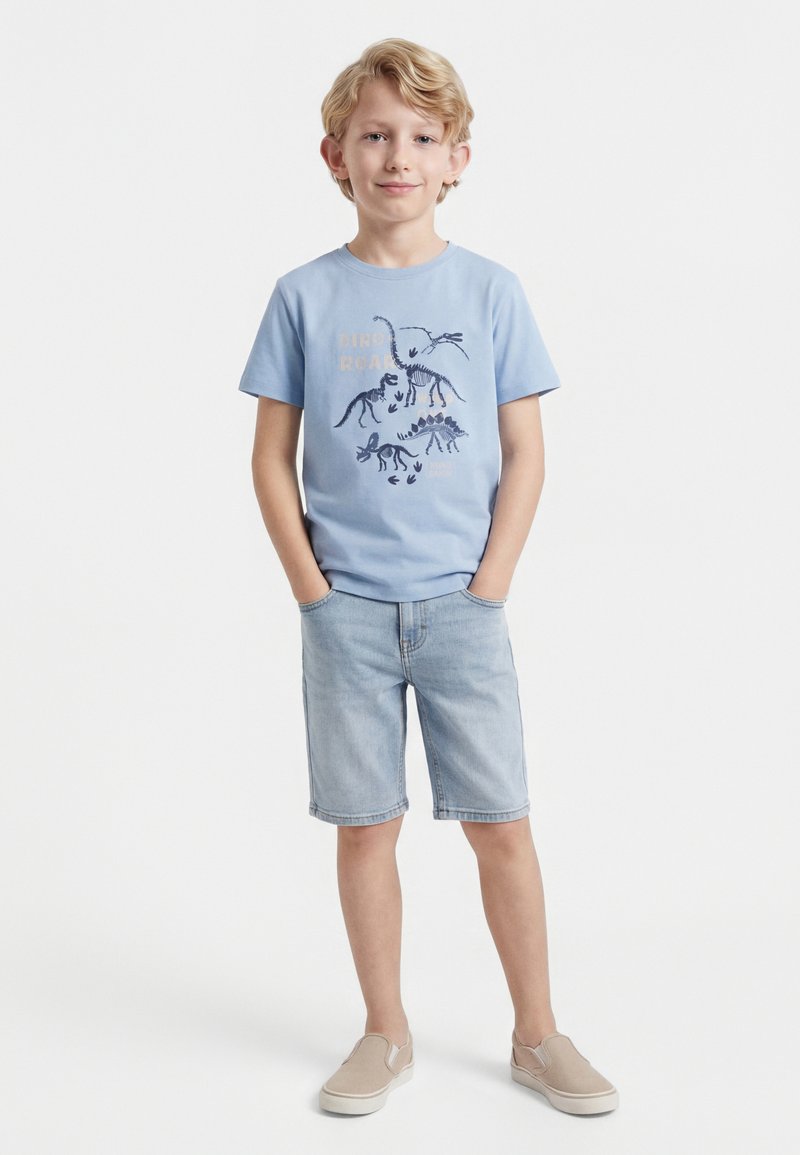 Ξανθός αγόρι με ανοιχτό μπλε μπλουζάκι με τύπωμα σκελετού δεινόσαυρου, ανοιχτόχρωμα τζιν σορτς και μπεζ παπούτσια χωρίς κορδόνια, στέκεται με τα χέρια στις τσέπες.