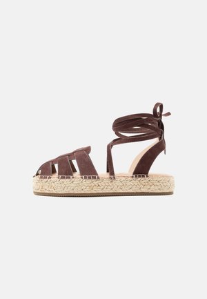 Brun ruskinds espadrille sandal med ankelremme og flettet jute-platformsål på hvid baggrund.