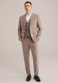 Hellbrauner Dreiteiler mit slim fit, bestehend aus einem einreiher Jacket, einer Weste mit Knöpfen und einer taillierten Hose. Schwarze Slipper.