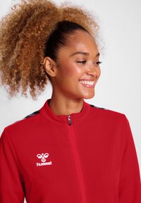 Rote Sportjacke mit hohem Kragen, Reißverschluss, schwarzen Akzenten an den Schultern und weißem Logo auf der Brust. Glatte Textur, sportlicher Schnitt.
