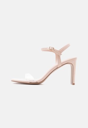 Beige High-Heel-Sandale mit dünnem Knöchelriemen und transparentem vorderem Riemen, geschlossene goldene Schnalle vor hellem neutralem Hintergrund.
