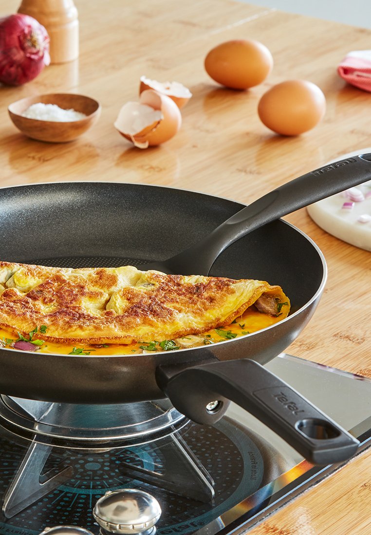 Omlet smażący się na czarnej patelni na gazowej kuchence, z jajkami, cebulą, solą i łopatką na drewnianym blacie.