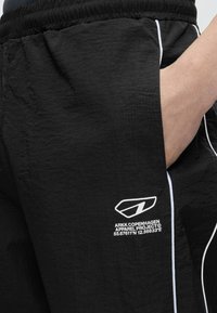 Κοντινή λήψη μαύρων αθλητικών σορτς με λευκές ραφές, ελαστική μέση, ένα χέρι στην τσέπη και λευκό λογότυπο στο μηρό.