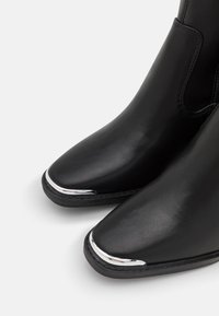 Botas de tobillo de cuero negro con punta afilada y acento cromado en la tapa del dedo. Textura suave con detalles cosidos alrededor de la suela.
