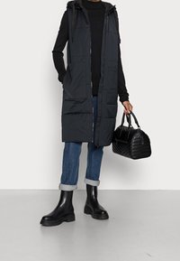 Długa czarna pikowana kamizelka z kapturem, zamkiem błyskawicznym i przednimi kieszeniami. W zestawie z ciemnymi dżinsami i czarnymi botkami, trzyma pikowaną torebkę.