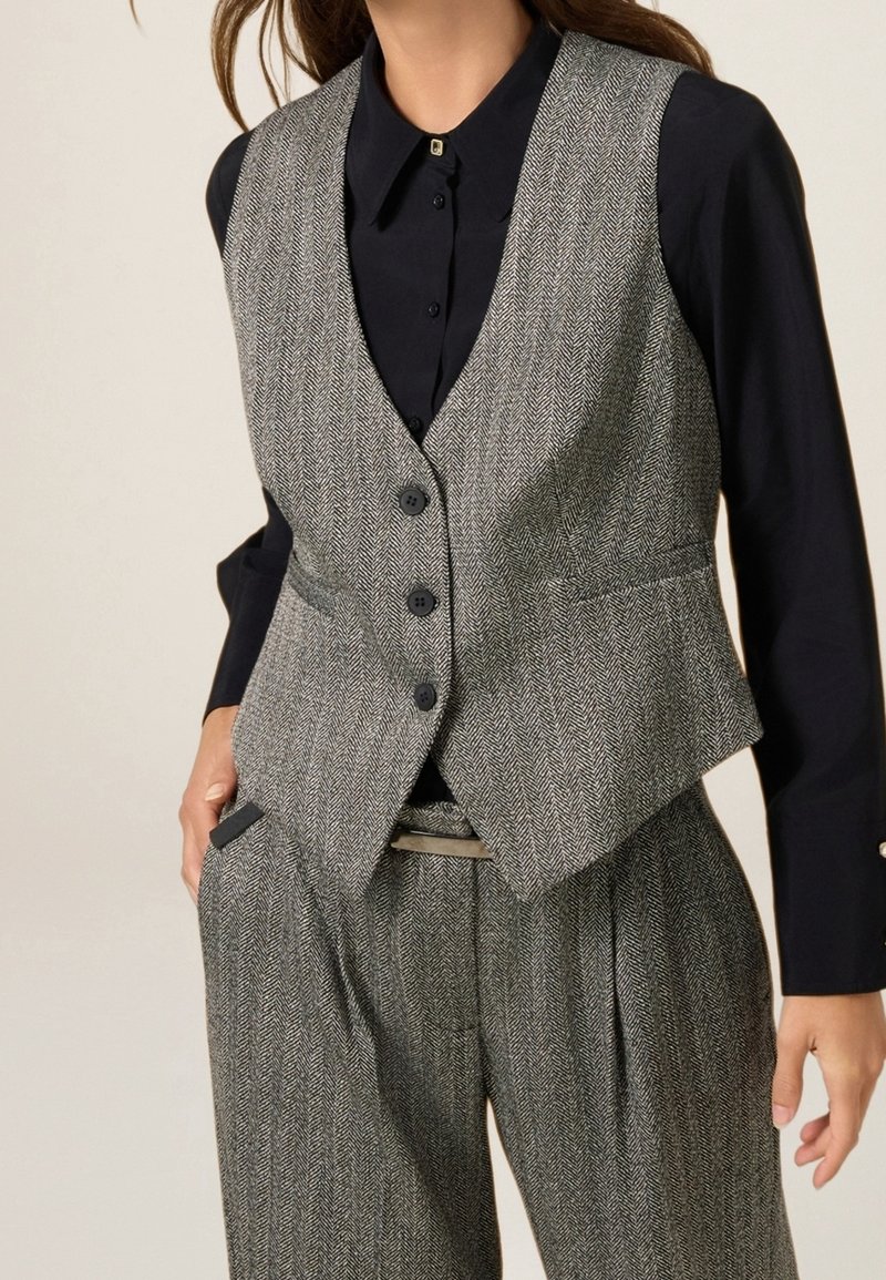 Femme portant un gilet en chevrons gris et un pantalon assorti sur une chemise noire à manches longues boutonnée avec une épingle de col carrée.