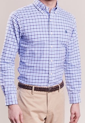 Mężczyzna w niebiesko-białej koszuli w kratę, wpuszczonej w beżowe spodnie z brązowym plecionym paskiem na jednolitym tle.