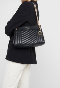 Donna che indossa un blazer nero e pantaloni bianchi, mentre porta una borsa a spalla in pelle trapuntata nera con tracolla in catena dorata e charm con logo.