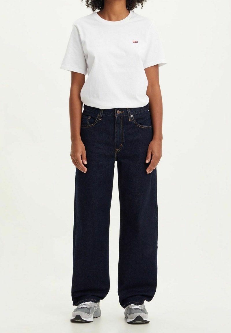 Camiseta blanca de manga corta con un pequeño logo rojo, combinada con jeans de mezclilla de pierna ancha en azul oscuro. Zapatillas grises con detalles blancos.