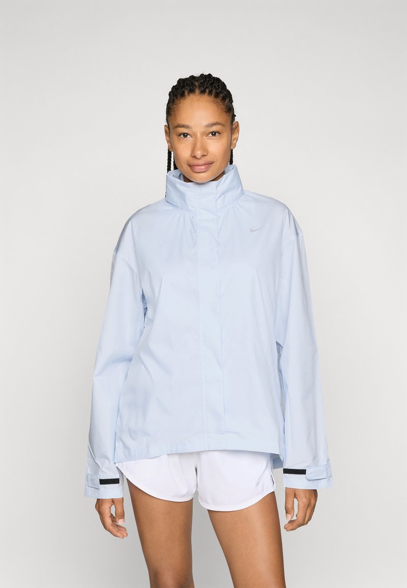 Chaqueta cortavientos azul claro con cuello alto y mangas largas. Presenta un diseño elegante y el logo de Nike en el pecho, combinada con pantalones cortos blancos.