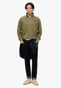 Olijfgroene polo met lange mouwen en kraag, witte onderhemd, marineblauwe broek en beige sneakers. Geruite flanellen stof om de taille geknoopt.