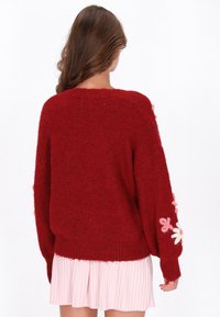 Röd stickad tröja med avslappnad passform, prydd med broderade rosa och vita blommor på ärmarna. Mjuk och fluffig textur.