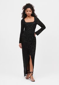 Abito nero in paillettes con maniche lunghe, scollo quadrato e uno spacco laterale. Tessuto morbido e strutturato con una silhouette aderente.