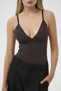 Donna che indossa un top fitted senza maniche di colore marrone scuro con spalline sottili e pantaloni neri larghi, con una mano in tasca, su uno sfondo chiaro e semplice.