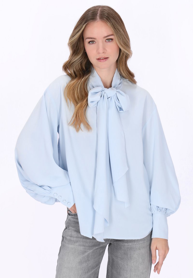 Fiatal nő hosszú, hullámos hajjal, világoskék blúzt visel masnis nyakkivágással és fodros ujjakkal, szürke farmerrel párosítva.