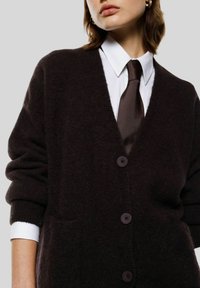 Cardigan marrone scuro con scollo a V, vestibilità oversize e tessuto morbido. Presenta grandi bottoni viola e maniche lunghe con polsini a costine.