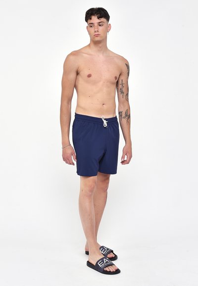 Costumi da bagno blu navy con cintura elastica e coulisse, realizzati in materiale leggero. Abbinati a ciabatte nere con scritte bianche.