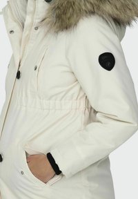 Parka bianca con cappuccio in finto pelo, accenti neri, vita elasticizzata e tasche laterali. Presenta bottoni e una toppa con logo sulla manica.