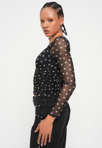 Schwarzes langärmliges Oberteil aus transparentem Pünktchenstoff, mit Raffung und figurbetontem Schnitt, kombiniert mit schwarzen Jeans mit Nietenverzierungen.