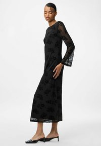 Zwarte bloemen kant jurk met lange, doorzichtige mouwen, een getailleerd lijfje en een vloeiende rok. Glanzende zwarte hakken mules complementeren de look.