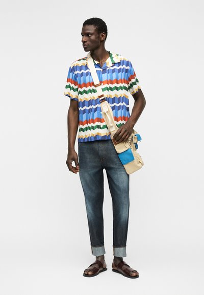 Άνδρας που φοράει πολύχρωμο πουκάμισο με ζιγκ ζαγκ μοτίβο, τζιν με ρεβέρ, καφέ σανδάλια και μια μπεζ τσάντα χιαστί με μπλε τσέπες.