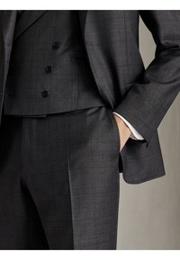 Homme portant un costume à carreaux gris foncé avec un gilet assorti, main dans la poche du pantalon, montrant la texture du tissu du costume et la coupe ajustée.