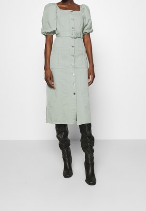 Mujer con vestido midi verde claro abotonado, con mangas abullonadas y cinturón, combinado con botas negras de caña alta y estilo holgado.