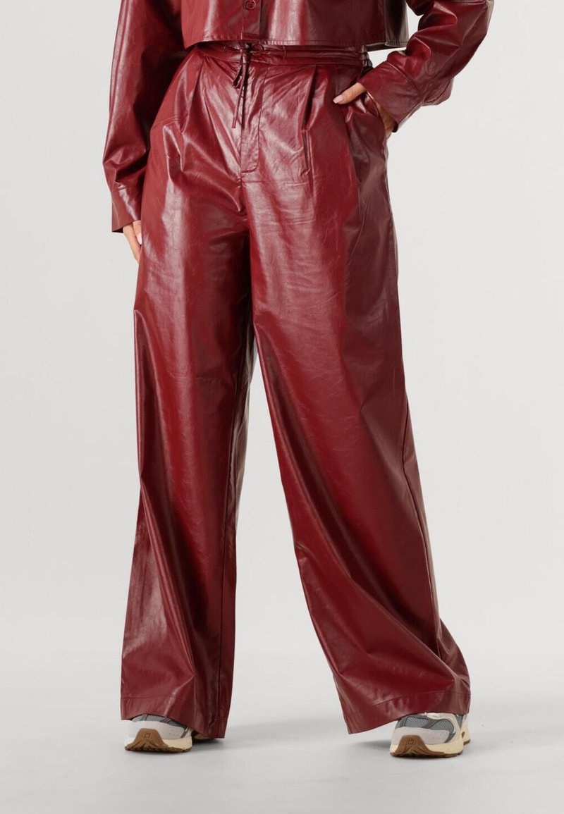 Persoon draagt hoge taille wijde rode leren broek en sneakers, met één hand in de zak, staand tegen een effen achtergrond.