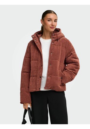 Femme portant une doudoune en velours côtelé couleur rouille avec capuche, chemise blanche, pantalon noir, et tenant un sac à main noir sur un fond uni.