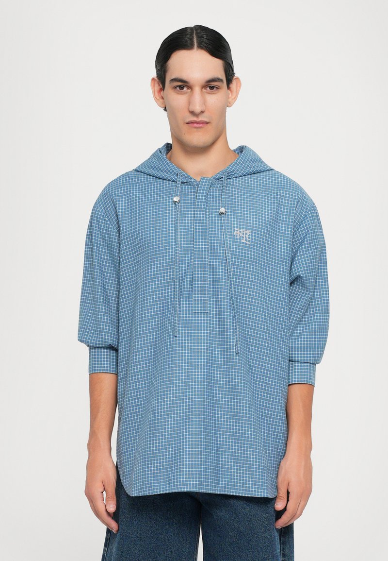 Camisa con capucha a cuadros azules, de corte holgado, con cierre de cordón y mangas cortas. La tela parece ligera y texturizada.