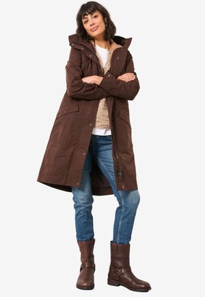 Frau mit kurzen dunklen Haaren, die einen langen braunen Mantel, einen beigen Pullover, blaue Jeans und braune Stiefeletten trägt, steht mit verschränkten Armen.
