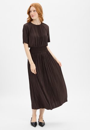 Brown pleated dress with short sleeves and a smocked waist, featuring a flowy skirt and subtle sheen. Worn with black pointed-toe shoes.