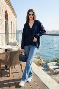 Frau trägt marineblauen Cardigan mit kleinen roten Herzen, blaue Jeans, weiße Sneakers und Sonnenbrille, steht auf einer Terrasse am Meer mit einer blauen Handtasche.