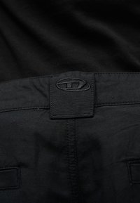 Κοντινό πλάνο της μέσης ενός μαύρου παντελονιού με ραμμένη θηλιά για ζώνη και μικρό ορθογώνιο λογότυπο πάνω από τη μεσαία ραφή.