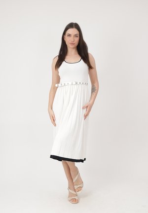 Kobieta z długimi ciemnymi włosami, ubrana w białą sukienkę bez rękawów z czarną lamówką i białym paskiem, stojąca z jedną ręką w kieszeni.