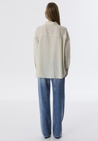 Lichtbeige oversized shirt met een subtiele textuur en een achterste schouderstuk, gecombineerd met blauwe jeans met verspreide witte spikkels.
