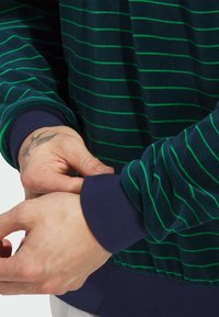Sweatshirt bleu marine rayé avec des stripes vertes, tissu texturé et poignets côtelés bleu foncé. Des mains ajustent la manche au niveau du poignet.