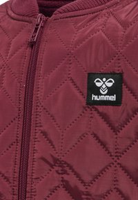 Burgundowa pikowana kurtka z zamkiem błyskawicznym z przodu i żebrowanym kołnierzem. Posiada wyraźnie widoczny czarny heksagonalny logo naszywkę.