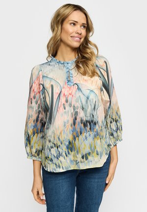 Kvinne med bølget blondt hår iført en løs, flerfarget bluse med abstrakt mønster og blå jeans, smilende lett og ser mot høyre.