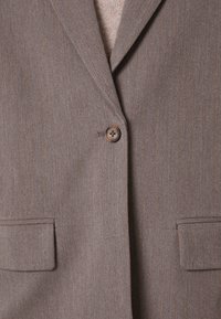 Blazer marron à fines rayures, col à revers cranté, fermeture par un bouton marron unique et deux poches avant à rabat sur un pull beige en maille.
