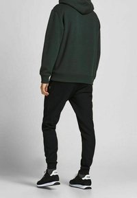 Donkergroene hoodie met een capuchon, gecombineerd met zwarte joggingsbroek en zwarte sneakers met witte accenten. De stof lijkt zacht en gestructureerd.