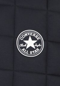 Svart, quiltet stoff har en rund hvit lapp med en stjerne, teksten "CONVERSE" og "Chuck Taylor" detaljer.