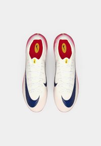 Un paio di scarpe da ginnastica bianche con loghi swoosh blu, lacci bianchi e solette rosse con loghi gialli e neri, viste dall'alto.