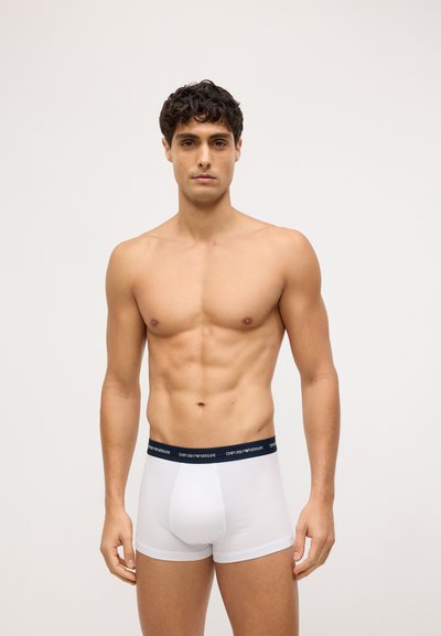 Baltie bokseri ar melnu elastību, uz kuras redzams "Emporio Armani" logotips. Gluda auduma un pieguļoša dizaina. Modelis demonstrē pilnu priekšējo skatu.