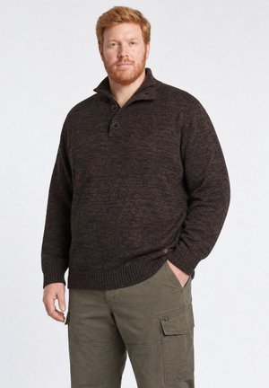 Mann mit rotem Haar, der einen dunkelbraunen Pullover und olivgrüne Cargohosen trägt, steht mit einer Hand in der Tasche.